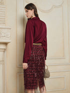 A woman, indoors against a decorative wall, wears the Social Queen 100% Silk Satin Wrap V-Neck Blouse with long sleeves and a sequined burgundy fringe skirt, holding a small gold handbag.