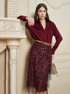 A woman stands indoors wearing the Social Queen 100% Silk Satin Wrap V-Neck Blouse and a sequined burgundy skirt, holding a small handbag while leaning against a cream-colored mantel.