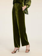 A person models Social Queen’s 100% Silk Velvet Wide-Leg Pants in vintage-inspired olive green, styled with metallic high-heeled sandals and a hand in their pocket, posed against a plain background.
