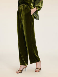 A person models Social Queen’s 100% Silk Velvet Wide-Leg Pants in vintage-inspired olive green, styled with metallic high-heeled sandals and a hand in their pocket, posed against a plain background.