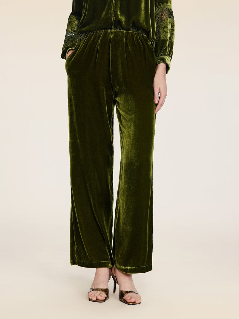A person stands against a plain background wearing Social Queen’s 100% Silk Velvet Wide-Leg Pants in olive green with brown strappy heels, hands casually in their pockets.