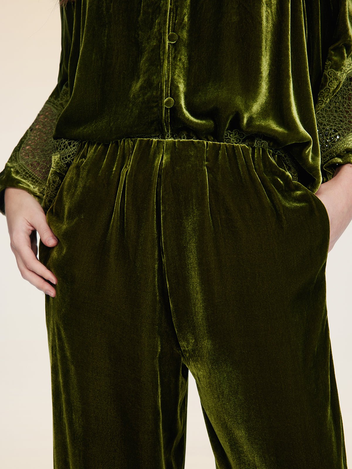 A person stands with hands in pockets, wearing vintage-inspired silk velvet wide-leg pants by Social Queen and a green top with lace-detailed sleeves.