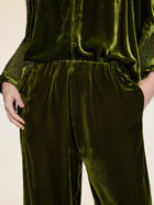 A person stands with hands in pockets, wearing vintage-inspired silk velvet wide-leg pants by Social Queen and a green top with lace-detailed sleeves.
