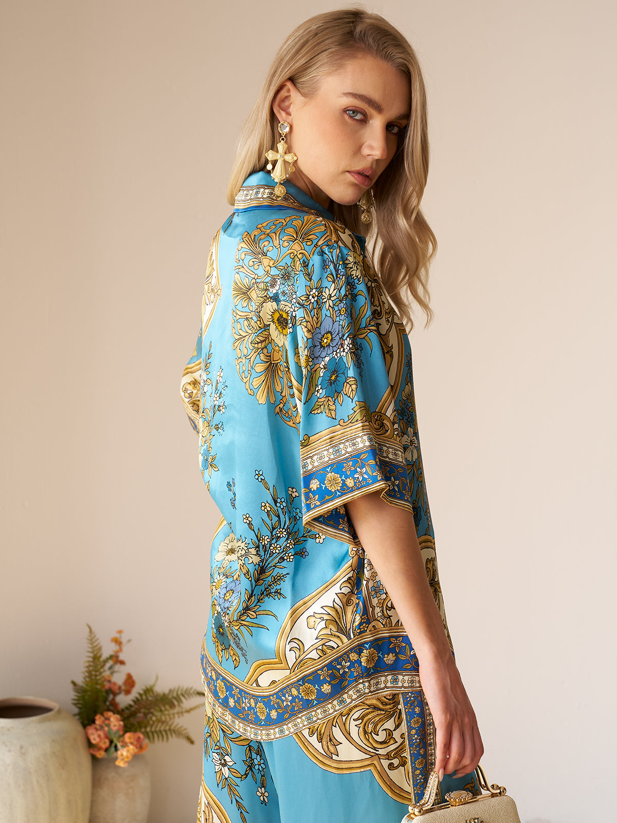 A woman with long blonde hair wears the Social Queen Baroque Engineered-Print Silk Shirt, featuring blue and gold hues, as she stands sideways looking over her shoulder with a vase of flowers in the background.