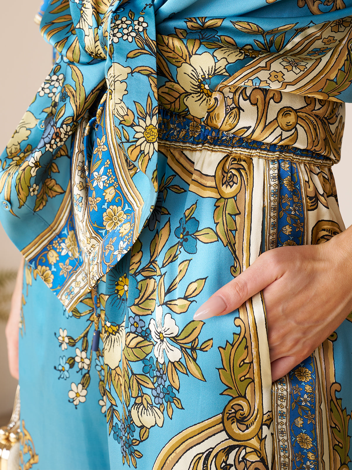 Close-up of a person wearing Social Queen's Baroque Engineered-Print Silk Wide-Leg Pants, featuring blue and gold floral and ornate patterns, with a hand resting in the side pocket.