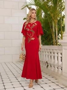 A woman wears the Social Queen Hand-Beaded Floral Mermaid Dress, featuring a long red design with chiffon cape sleeves, as she stands on a tiled patio beside a white railing and lush green plants.