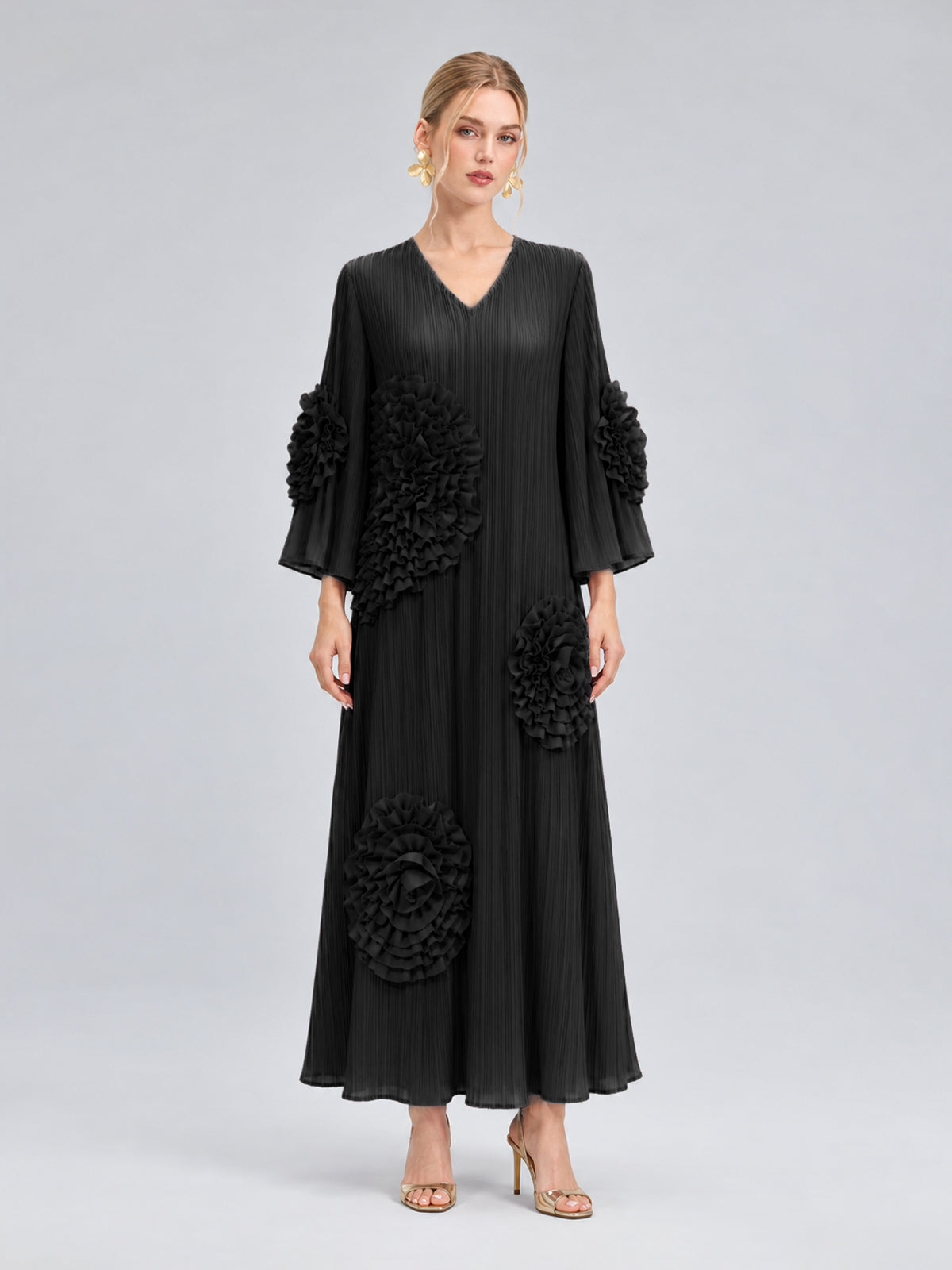 A woman stands against a plain grey background wearing the Social Queen 3D Floral Pleated Dress—a long, black maxi dress featuring large floral appliqué embellishments—paired with gold earrings and gold heels.