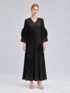 A woman stands against a plain grey background wearing the Social Queen 3D Floral Pleated Dress—a long, black maxi dress featuring large floral appliqué embellishments—paired with gold earrings and gold heels.