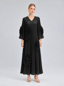 A woman stands against a plain grey background wearing the Social Queen 3D Floral Pleated Dress—a long, black maxi dress featuring large floral appliqué embellishments—paired with gold earrings and gold heels.