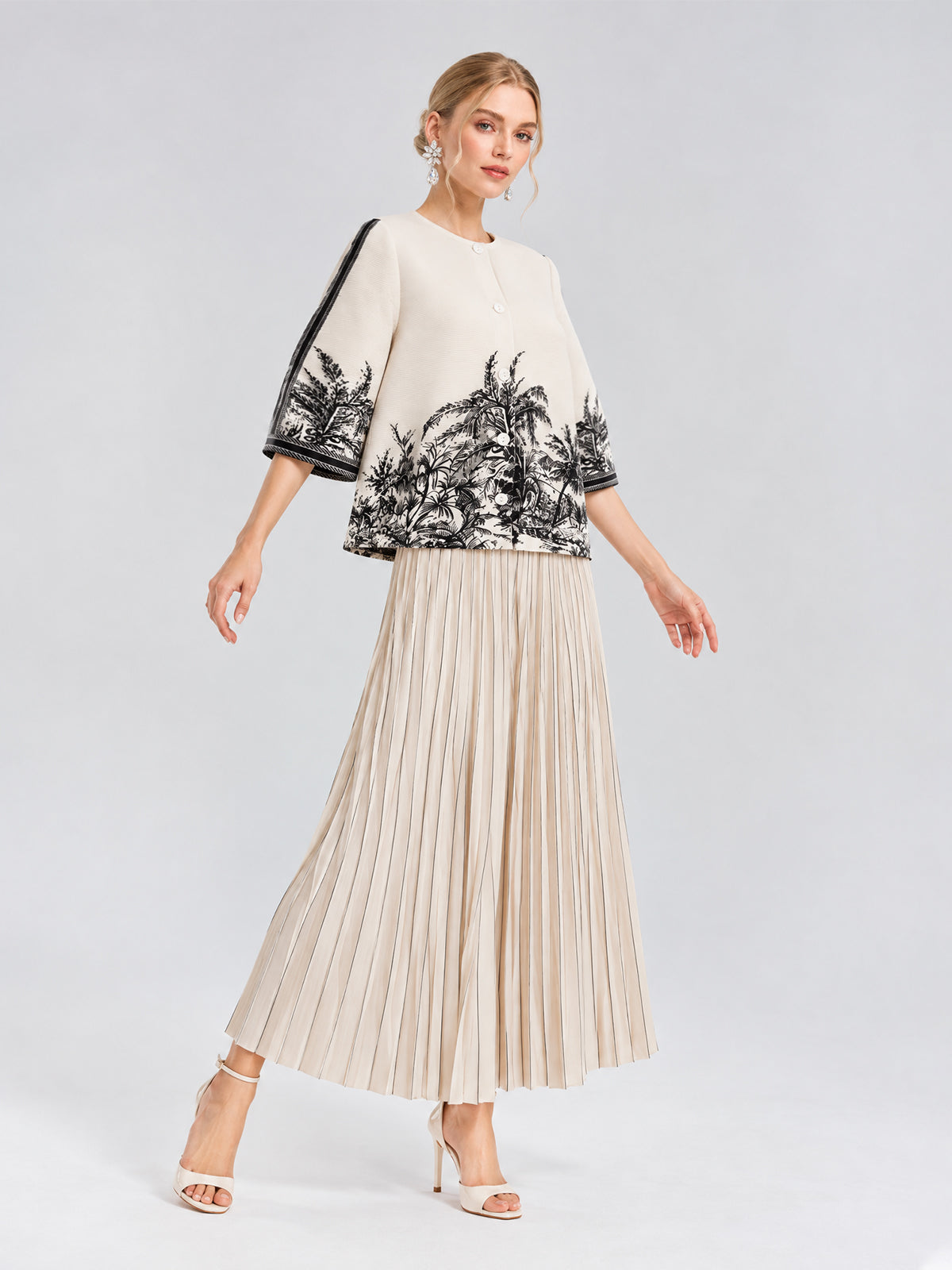 A woman stands against a plain background wearing the Social Queen Palm Tree Print Pleated Blouse with a cropped silhouette, styled with a beige pleated skirt and white heeled sandals.