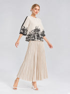 A woman stands against a plain background wearing the Social Queen Palm Tree Print Pleated Blouse with a cropped silhouette, styled with a beige pleated skirt and white heeled sandals.