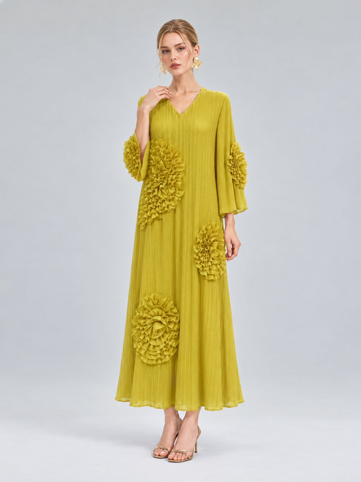 A woman models the Social Queen 3D Floral Pleated Dress—a long, pleated yellow maxi adorned with large floral appliqués on the sleeves and skirt—standing against a plain light gray background.