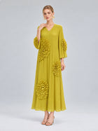 A woman models the Social Queen 3D Floral Pleated Dress—a long, pleated yellow maxi adorned with large floral appliqués on the sleeves and skirt—standing against a plain light gray background.