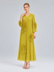 A woman models the Social Queen 3D Floral Pleated Dress—a long, pleated yellow maxi adorned with large floral appliqués on the sleeves and skirt—standing against a plain light gray background.