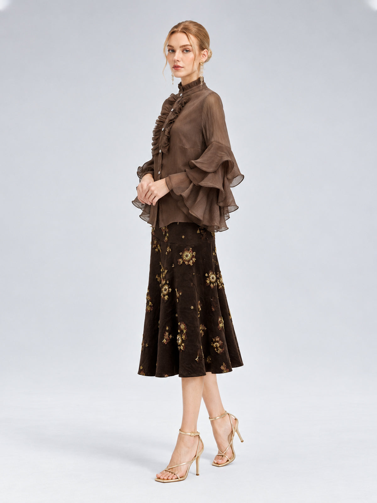 A woman is wearing a sheer brown ruffled blouse and the Floral Beaded Midi Skirt by Social Queen, featuring an elegant A-line silhouette, paired with strappy gold high heels, standing against a plain light background.