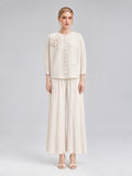 A woman stands against a plain background wearing the Social Queen 3D Floral Pleated Two-Piece Set in cream-white, which features a floral appliqué top and wide-leg pleated pants paired with open-toed heels.
