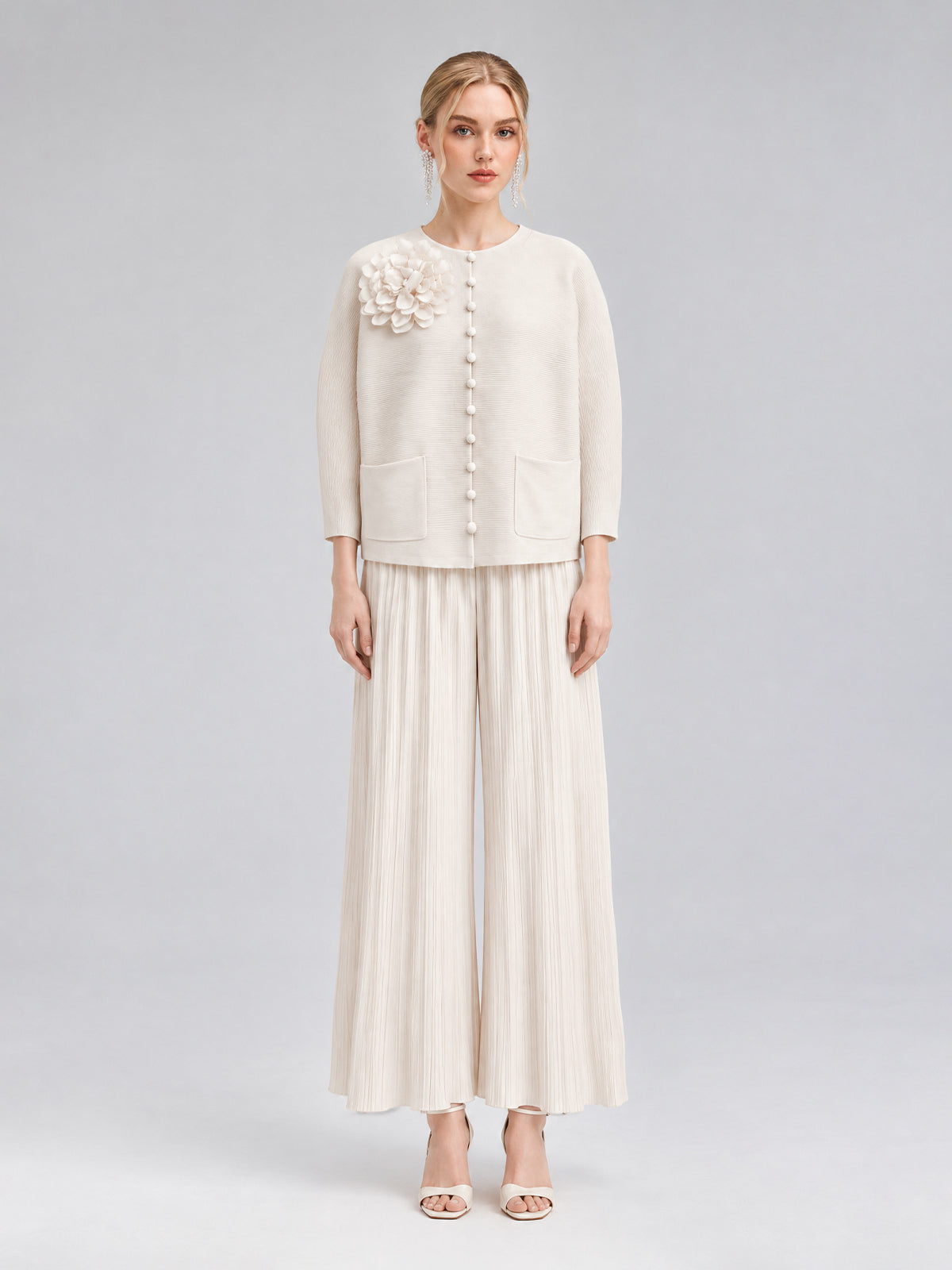 A woman stands against a plain background wearing the Social Queen 3D Floral Pleated Two-Piece Set in cream-white, which features a floral appliqué top and wide-leg pleated pants paired with open-toed heels.