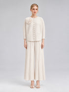 A woman stands against a plain background wearing the Social Queen 3D Floral Pleated Two-Piece Set in cream-white, which features a floral appliqué top and wide-leg pleated pants paired with open-toed heels.