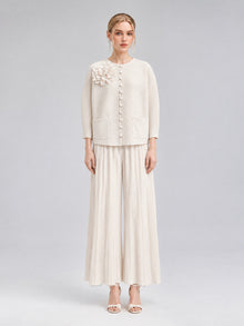 A woman stands against a plain background wearing the Social Queen 3D Floral Pleated Two-Piece Set in cream-white, which features a floral appliqué top and wide-leg pleated pants paired with open-toed heels.