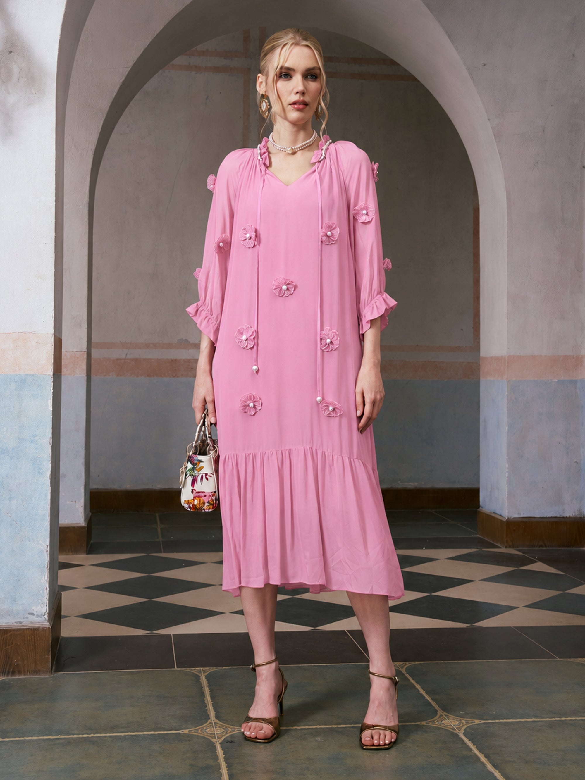 A woman stands indoors, posing elegantly in front of an arched doorway, wearing the Social Queen 3D Floral Pearl Mulberry Silk Dress in a loose-fitting pink silhouette, paired with strappy heels and a patterned handbag.