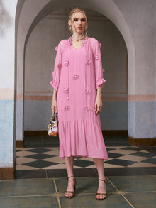 A woman stands indoors, posing elegantly in front of an arched doorway, wearing the Social Queen 3D Floral Pearl Mulberry Silk Dress in a loose-fitting pink silhouette, paired with strappy heels and a patterned handbag.