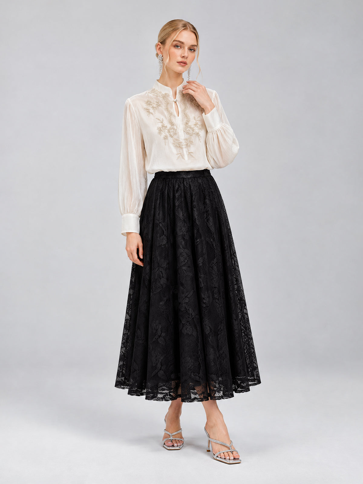 A woman stands against a plain gray background wearing the Social Queen Floral Vine Embroidered Blouse, paired with a long black lace skirt and silver sandals.