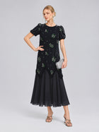 A woman stands against a plain background, wearing the Social Queen Beaded Floral Silk Velvet Dress in navy blue velvet and chiffon with intricate beaded floral embroidery. She completes the look with a silver clutch and heels.
