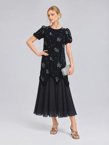 A woman stands against a plain background, wearing the Social Queen Beaded Floral Silk Velvet Dress in navy blue velvet and chiffon with intricate beaded floral embroidery. She completes the look with a silver clutch and heels.