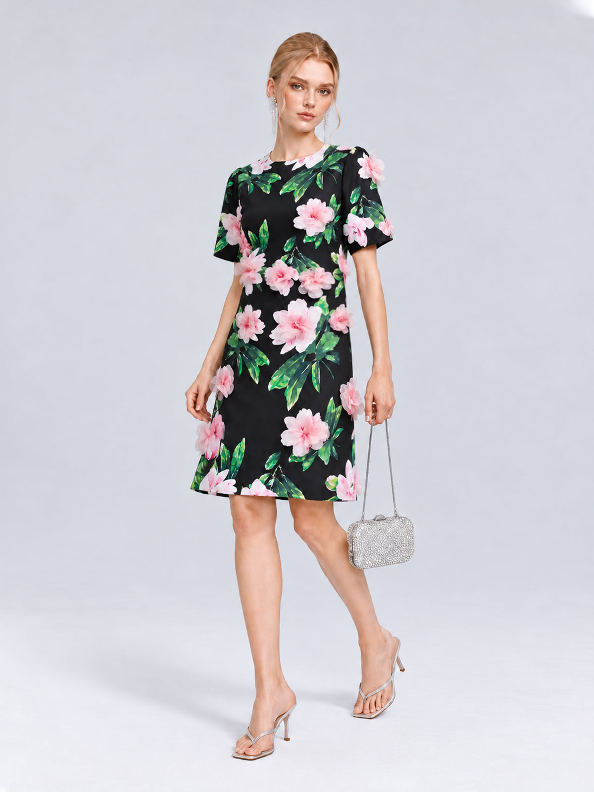 A woman stands against a plain light gray background wearing the Social Queen 3D Peony Floral Shift Dress paired with clear heeled sandals and carrying a small silver handbag.