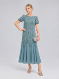 A woman wearing the Social Queen Beaded Floral Silk Velvet Dress—crafted in a light blue silk-blend velvet with beaded floral embroidery on the short sleeves—holds a silver clutch and stands against a plain background, making this the perfect dress for wedding occasions.