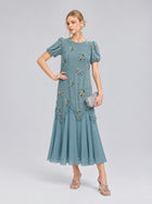 A woman wearing the Social Queen Beaded Floral Silk Velvet Dress—crafted in a light blue silk-blend velvet with beaded floral embroidery on the short sleeves—holds a silver clutch and stands against a plain background, making this the perfect dress for wedding occasions.