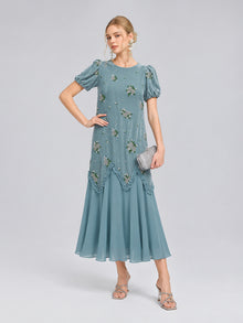 A woman wearing the Social Queen Beaded Floral Silk Velvet Dress—crafted in a light blue silk-blend velvet with beaded floral embroidery on the short sleeves—holds a silver clutch and stands against a plain background, making this the perfect dress for wedding occasions.