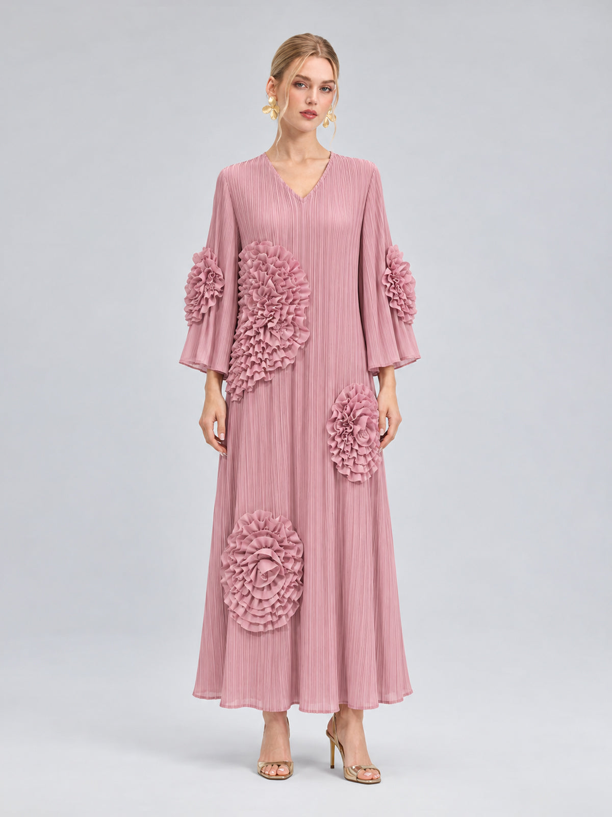 A woman stands against a plain gray background wearing the Social Queen 3D Floral Pleated Dress—a long pink maxi dress featuring large, textured floral appliqués—paired with gold heels.
