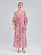 A woman stands against a plain gray background wearing the Social Queen 3D Floral Pleated Dress—a long pink maxi dress featuring large, textured floral appliqués—paired with gold heels.