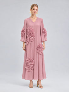 A woman stands against a plain gray background wearing the Social Queen 3D Floral Pleated Dress—a long pink maxi dress featuring large, textured floral appliqués—paired with gold heels.