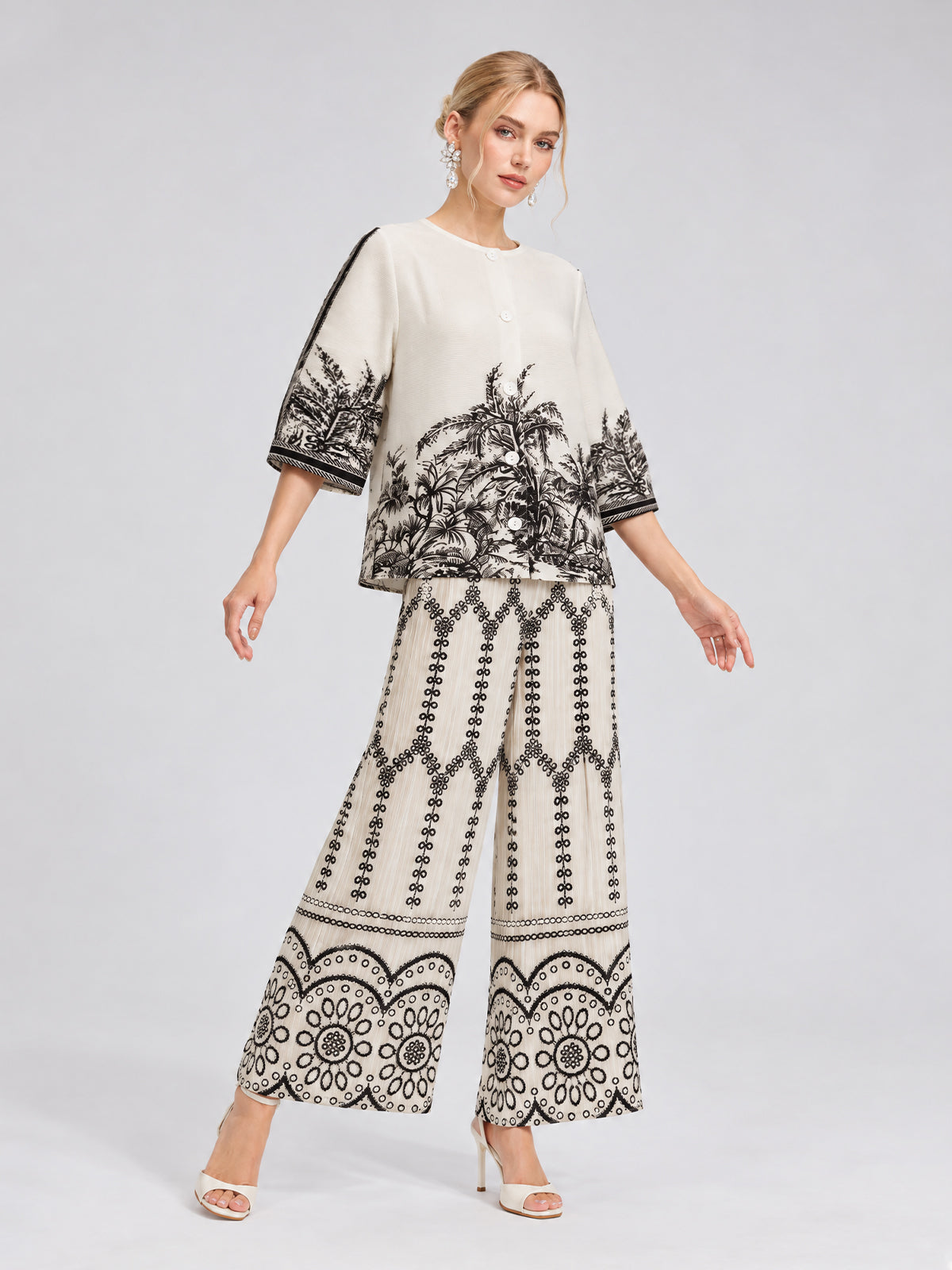 A woman stands against a plain light background wearing Social Queen’s Geometric Print Pleated Wide-Leg Pants, styled as part of a cream-colored matching set featuring black botanical and geometric patterns, paired with heeled sandals.