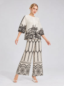 A woman stands against a plain light background wearing Social Queen’s Geometric Print Pleated Wide-Leg Pants, styled as part of a cream-colored matching set featuring black botanical and geometric patterns, paired with heeled sandals.