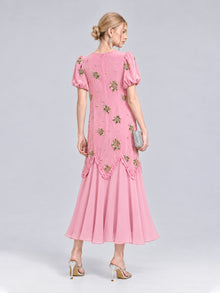 A woman stands with her back to the camera, wearing the Social Queen Beaded Floral Silk Velvet Dress—a pink, ankle-length, short-sleeve design crafted from a silk-blend velvet and detailed with intricate beaded floral embroidery and embellishments. She holds a silver clutch and wears heels.
