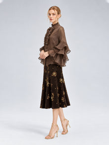A woman stands in profile against a plain gray background, wearing the Social Queen Pleated Layered Ruffle Blouse—crafted from sheer brown polyester chiffon with pleated ruffles and a high neckline—paired with a dark floral midi skirt and light strappy high heels.