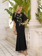 On a balcony, a woman wears the Social Queen Hand-Beaded Floral Mermaid Dress with chiffon cape sleeves, gold jewelry, and a textured gold clutch. Lush green plants fill the background.
