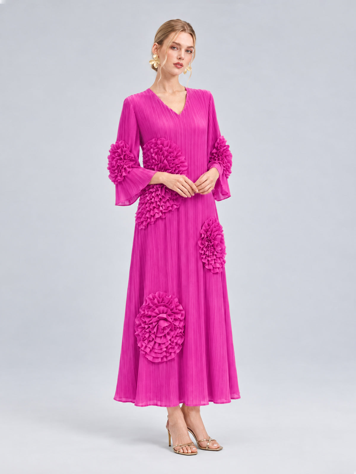 A woman models the Social Queen 3D Floral Pleated Dress—a long, vibrant pink maxi dress featuring large floral appliqué ruffle accents and precise pleating—paired with heeled sandals, posed against a plain light gray background.