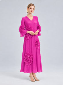 A woman models the Social Queen 3D Floral Pleated Dress—a long, vibrant pink maxi dress featuring large floral appliqué ruffle accents and precise pleating—paired with heeled sandals, posed against a plain light gray background.