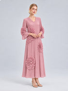 A woman wearing the Social Queen 3D Floral Pleated Dress—an elegant, long pink maxi dress featuring pleats and large, textured floral appliqué embellishments—stands against a plain light background.