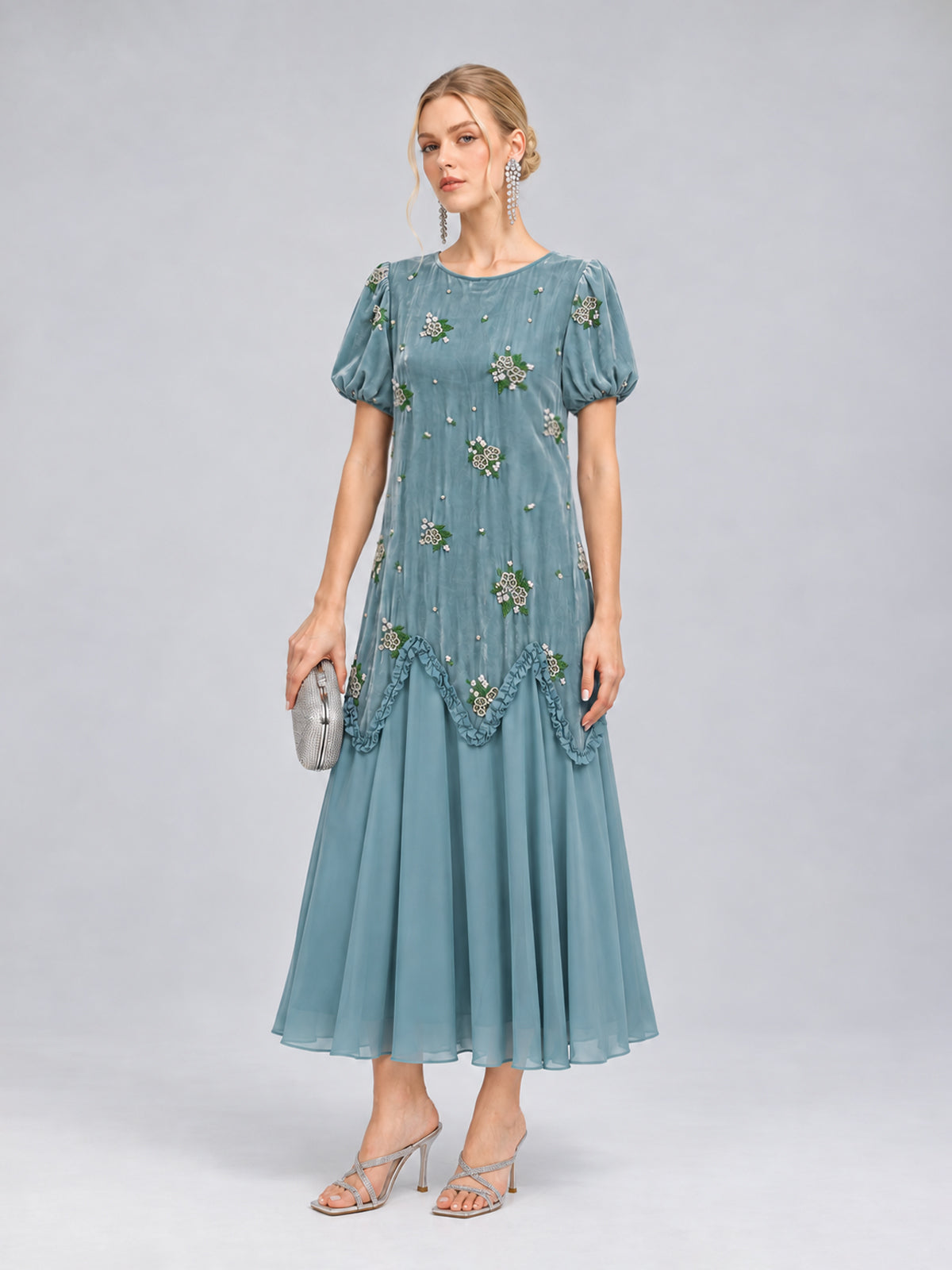 A woman stands against a plain gray background wearing the Social Queen Beaded Floral Silk Velvet Dress—a light blue midi dress with intricate beaded floral embroidery and puff sleeves. She completes the look with silver heels and a matching silver clutch, making it a perfect choice for a wedding dinner.
