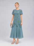 A woman stands against a plain gray background wearing the Social Queen Beaded Floral Silk Velvet Dress—a light blue midi dress with intricate beaded floral embroidery and puff sleeves. She completes the look with silver heels and a matching silver clutch, making it a perfect choice for a wedding dinner.