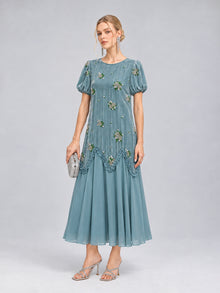 A woman stands against a plain gray background wearing the Social Queen Beaded Floral Silk Velvet Dress—a light blue midi dress with intricate beaded floral embroidery and puff sleeves. She completes the look with silver heels and a matching silver clutch, making it a perfect choice for a wedding dinner.