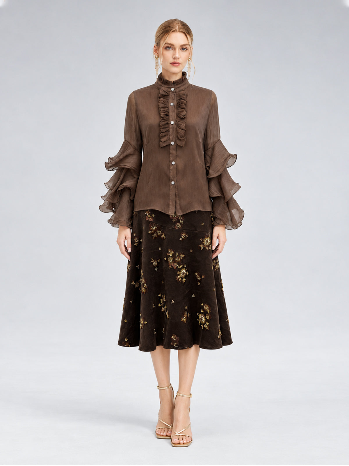 A woman stands facing forward, wearing the Pleated Layered Ruffle Blouse from Social Queen—a brown blouse with layered ruffle sleeves crafted from lightweight polyester chiffon—paired with a dark floral midi skirt and strappy beige heels against a neutral background.