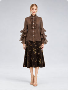 A woman stands facing forward, wearing the Pleated Layered Ruffle Blouse from Social Queen—a brown blouse with layered ruffle sleeves crafted from lightweight polyester chiffon—paired with a dark floral midi skirt and strappy beige heels against a neutral background.