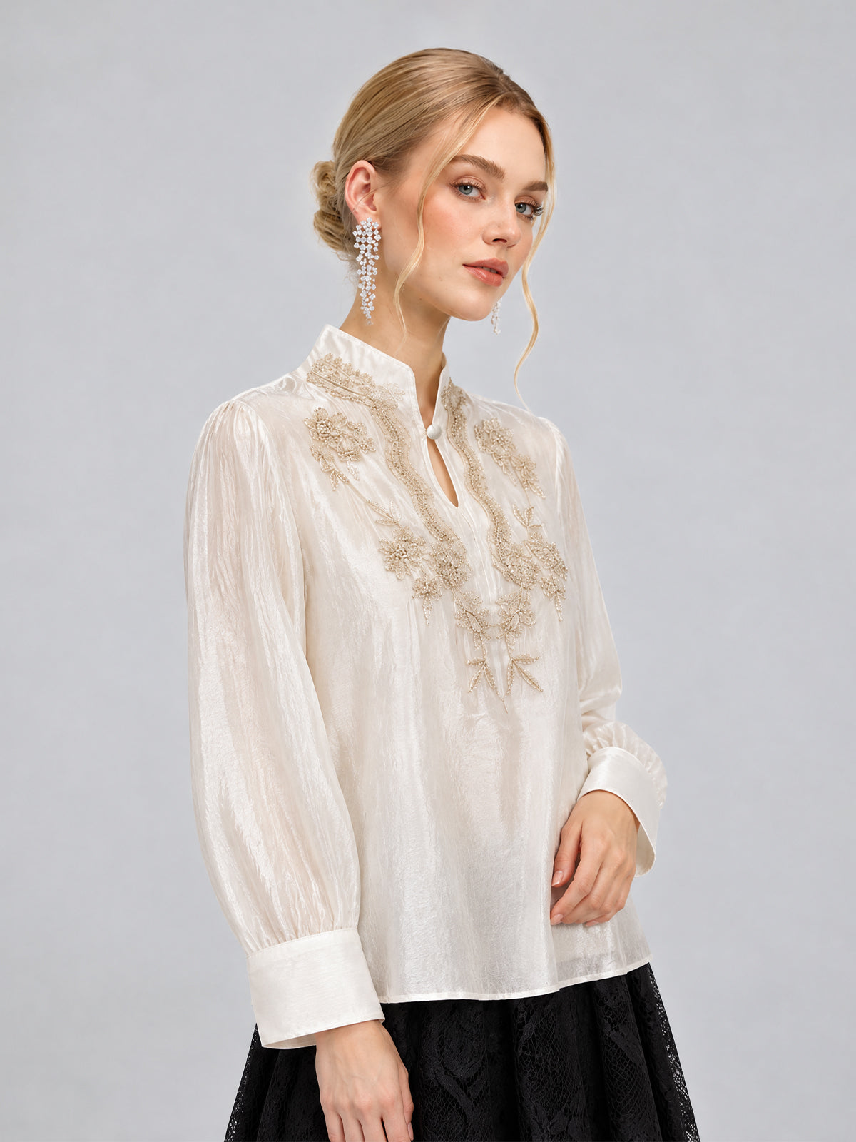 A woman wearing the Social Queen Floral Vine Embroidered Blouse in ivory, paired with a black skirt and long crystal earrings, stands against a plain gray background.