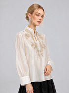 A woman wearing the Social Queen Floral Vine Embroidered Blouse in ivory, paired with a black skirt and long crystal earrings, stands against a plain gray background.